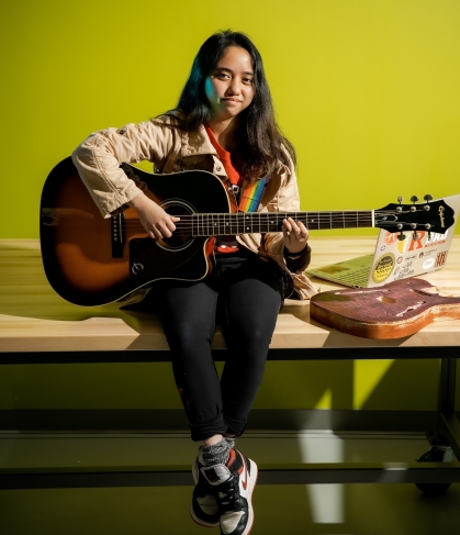 Young woman with long dark hair sits on table holding a guitar.