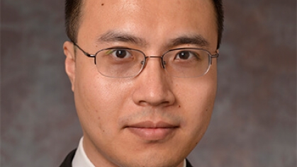 Head shot of Asian male with short black hair and eyeglasses wearing a black suit, white tie, and a bllue patterned tie.
