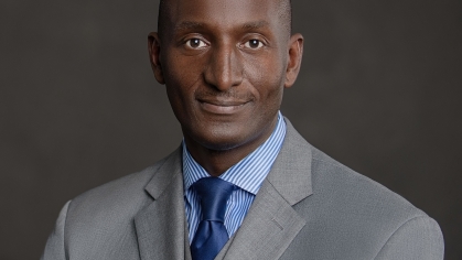 Black male head shot wearing a gray suit with light blue shirt and dark blue tie.