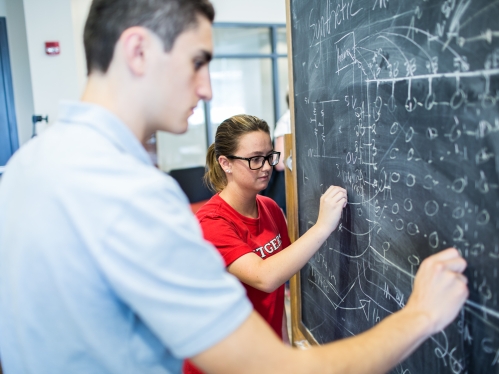 Two students writing formulas on greenboard