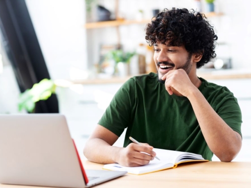 Young man with black curly hair and a beard, wearing a dark green shirt, smiles will looking at a laptop.